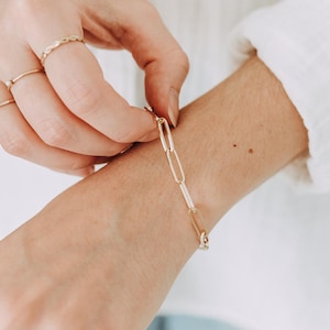 May include: A gold link bracelet is being fastened on a wrist. The bracelet has elongated oval links. Several gold rings are on the fingers. The background is a soft, neutral colour.