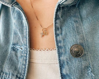 May include: A gold necklace with a small cross pendant and a letter "B" pendant. The necklace is worn over a ribbed, cream-colored tank top and a light blue denim jacket.