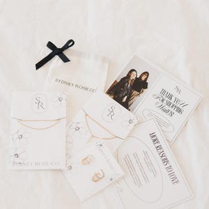 May include: An assortment of jewelry and packaging from Sydney Rose Co. includes necklaces, earrings, and cards. The cards feature the text "Thank You For Shopping With Us!" and "More Reasons to Love." A black ribbon and a small white bag are also present.
