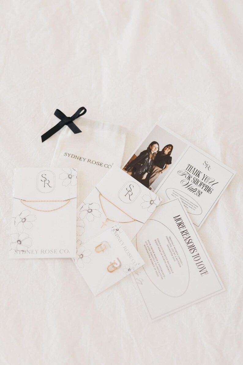 May include: An assortment of jewelry and packaging from Sydney Rose Co. includes necklaces, earrings, and cards. The cards feature the text "Thank You For Shopping With Us!" and "More Reasons to Love." A black ribbon and a small white bag are also present.