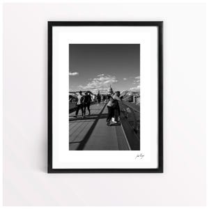 May include: Black and white framed photograph of a bridge with people walking on it. The background features a building with a dome and a clear sky with clouds. The photograph is signed in the bottom right corner.