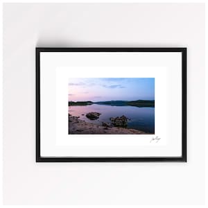 May include: A framed landscape photograph featuring a calm lake at twilight with soft pink and blue hues in the sky, dark rocky formations along the shoreline, and a signature in the bottom right corner of the white matting inside the black frame.