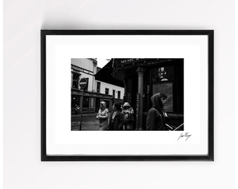 May include: A black and white framed photograph depicting a street scene with three individuals wearing hooded jackets standing near a building marked with the word MUSIC. A circular traffic sign is visible on the left side of the frame.