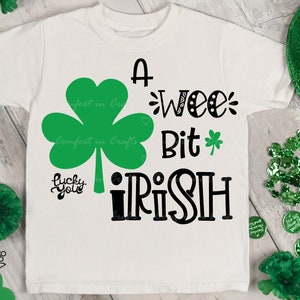 May include: A white t-shirt with a green shamrock and the text "A wee bit Irish" in black. The shirt is surrounded by green shamrocks, green glitter, and a green hat.
