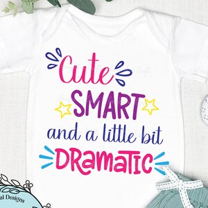 Könnte beinhalten: Weißer Baby-Strampler mit dem Text "Cute SMART and a little bit DRAMATIC" in bunten Buchstaben. Der Strampler ist von Herzen und einer Babyrassel umgeben.