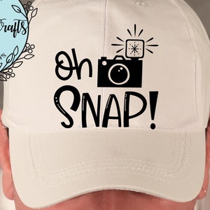 May include: White baseball cap with black text that reads "oh snap!" and a black silhouette of a camera with a flash.