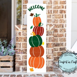 May include: A white wooden sign with the text "Welcome" and a stack of four pumpkins in orange, green, and red. The pumpkins are stacked on top of each other and have a fall leaf design. The sign is decorated with a wreath of leaves and the text "Comfort in Crafts" in a circle.