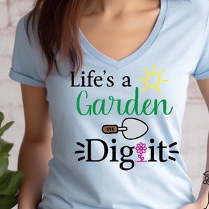 Op de afbeelding: Een lichtblauw t-shirt met een v-hals en de tekst "Life's a Garden Dig it" in zwarte en groene letters. Een kleine zon en een tuinschep zijn ook in het ontwerp opgenomen.