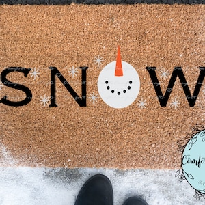 May include: A rectangular doormat with the word "SNOW" in black letters. The "O" is replaced with a snowman face. Snowflakes are scattered around the letters. The mat is brown and sits on a snowy surface.