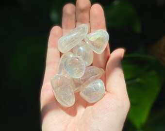 May include: A collection of iridescent clear quartz tumbled stones in a hand. The stones are smooth and have a rainbow sheen.