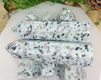 May include: A group of six white and black speckled crystal points, also known as leopard skin jasper, arranged in a stack on a white surface.