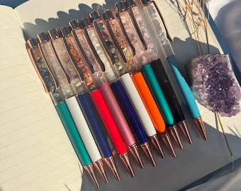 May include: A selection of pens with rose gold detailing. The pens have clear barrels filled with small stones and feature various coloured bases, including teal, white, red, and black. They are arranged on an open notebook.
