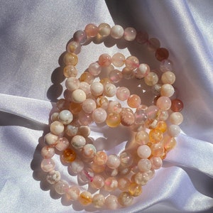 Puede incluir: Un conjunto de tres pulseras hechas con cuentas de ágata rosa y blanca. Las cuentas son redondas y tienen un acabado natural y pulido.