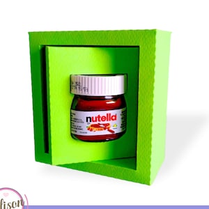Peut inclure: Une boîte en carton verte avec une découpe révélant un petit pot en verre de Nutella. Le pot a un couvercle blanc et une étiquette rouge avec le mot "Nutella" imprimé en blanc.