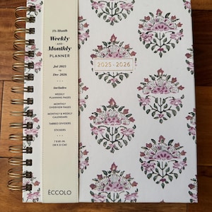 May include: A 15-month weekly and monthly planner with a floral pattern in pink and green on a white background. The planner includes weekly planning pages, monthly calendars, and tabbed dividers. The size is 7 x 8½ inches.