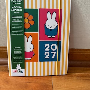 May include: A 17-month monthly planner with a yellow and white striped cover featuring Miffy illustrations, a green vertical band with text, and a yellow elastic closure. The cover displays a flower, Miffy in a blue outfit, Miffy in a red shirt, and the year 2027.