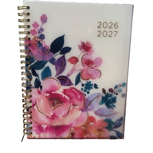 May include: A spiral-bound planner with a white cover featuring a watercolor floral design in shades of pink, purple, and blue. The front cover displays the years 2026 and 2027 in gold-colored text. The binding consists of a gold-toned metal coil.
