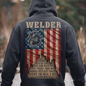 May include: Black hooded sweatshirt with a distressed American flag design and the text "WELDER" above the flag. The flag has a welding helmet and welding torch in the top left corner. The text below the flag reads "LORD, BLESS THIS JOB AND KEEP ME SAFE TODAY. MAY MY HANDS REFLECT YOUR LOVE IN ALL I DO ★★★I PRAY★★★".