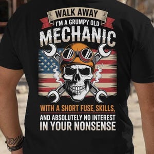 May include: Black t-shirt with a graphic of a skull wearing a mechanic's hat and goggles, with crossed wrenches and an American flag. The text reads "Walk Away I'm a Grumpy Old Mechanic With a Short Fuse, Skills, and Absolutely No Interest in Your Nonsense."