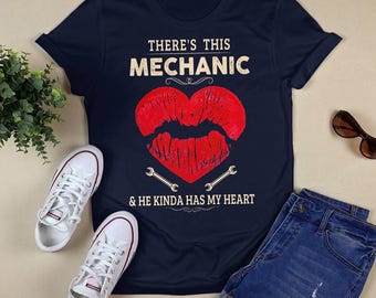 Puede incluir: Una camiseta azul marino con el texto "There's this Mechanic & he kinda has my heart". Un corazón rojo con una impresión de lápiz labial está en el centro de la camiseta. Dos símbolos de llave inglesa están debajo del corazón.