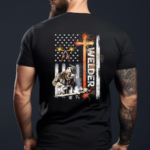 May include: Black t-shirt with a distressed American flag design featuring a welder working with a welding torch and the word "Welder" written vertically. A cross is behind the welder.