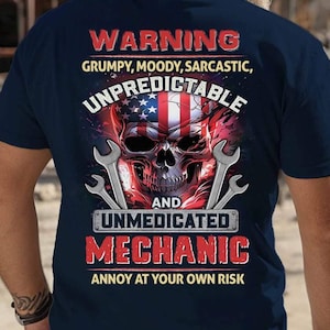 Könnte beinhalten: Ein marineblaues T-Shirt mit einem Totenkopf-Motiv und dem Text "WARNING GRUMPY, MOODY, SARCASTIC, UNPREDICTABLE AND UNMEDICATED MECHANIC ANNOY AT YOUR OWN RISK". Der Totenkopf trägt ein Bandana mit amerikanischer Flagge und hält zwei Schraubenschlüssel.