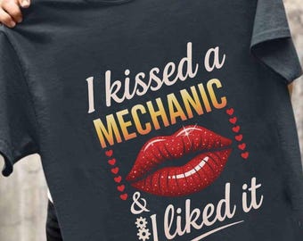 Puede incluir: Una camiseta gris oscuro con el texto "I kissed a MECHANIC & I liked it" en blanco y naranja. Una impresión de labios rojos cubiertos de purpurina está sobre el texto con pequeños corazones blancos alrededor.