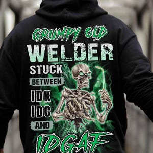 May include: Black hoodie with a green graphic of a skeleton holding a welding torch. The text on the hoodie reads "Grumpy Old Welder Stuck Between IDK IDC And IDGAF".