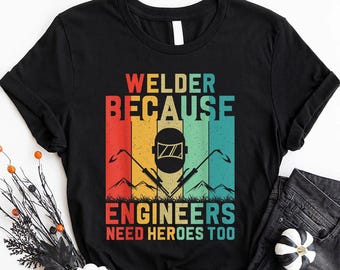 May include: Black t-shirt with a vintage-style graphic that says "Welder Because Engineers Need Heroes Too". The graphic features a welder's helmet and tools in a rainbow color scheme.