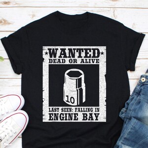 以下が含まれることがあります： WANTED DEAD OR ALIVEと書かれた白いグラフィックがプリントされた黒いTシャツ。ポスターには、10mmのソケットレンチと「LAST SEEN: FALLING IN ENGINE BAY」というテキストが描かれています。