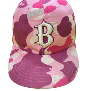 May include: Pink camouflage baseball cap with a white embroidered "B" on the front. The cap features a curved brim and a fabric strap for adjustment. The hat is made of a lightweight material.