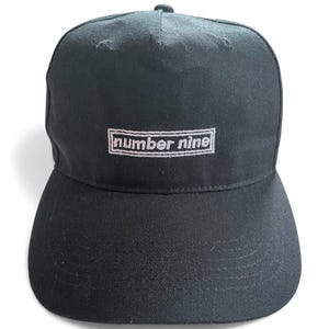 K&ouml;nnte beinhalten: Schwarze Baseballkappe mit gebogenem Schirm. Auf der Vorderseite der Kappe ist der Text "number nine" in Wei&szlig; in einem rechteckigen Rahmen gestickt. Die Kappe ist aus strapazierf&auml;higem Stoff gefertigt.