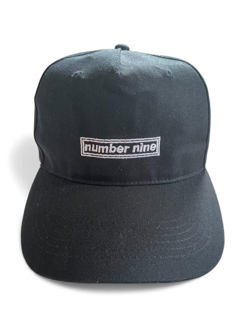 K&ouml;nnte beinhalten: Schwarze Baseballkappe mit gebogenem Schirm. Auf der Vorderseite der Kappe ist der Text "number nine" in Wei&szlig; in einem rechteckigen Rahmen gestickt. Die Kappe ist aus strapazierf&auml;higem Stoff gefertigt.