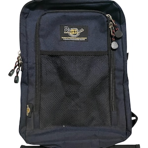 Puede incluir: Mochila azul marino oscuro con un bolsillo de malla negro y un compartimento frontal con cremallera. La mochila presenta el logotipo de Dr. Martens y el texto: TECHNICAL SPECIFICATIONS & STRENGTH. WORLDWIDE DESIGN.
