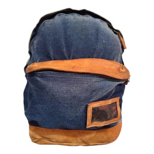 May include: A blue denim backpack featuring a large front pocket with a tan leather flap and a matching tan leather base. The front of the bag includes a rectangular clear plastic identification window framed in tan leather with a zipper closure.