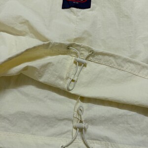 May include: A cream-colored windbreaker jacket featuring a dark blue rectangular patch with the white and red Fila Biella Italia logo. The garment includes two white elastic drawstrings with plastic toggles for adjustable cinching at the waist.