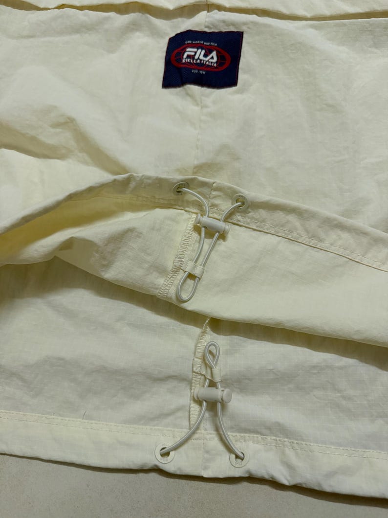 May include: A cream-colored windbreaker jacket featuring a dark blue rectangular patch with the white and red Fila Biella Italia logo. The garment includes two white elastic drawstrings with plastic toggles for adjustable cinching at the waist.