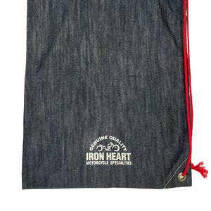 以下が含まれることがあります： 赤色の紐が付いた濃いグレーのデニム巾着袋。「GENUINE QUALITY IRON HEART MOTORCYCLE SPECIALTIES」の白文字が特徴です。紐を通すための補強されたハトメが付いています。