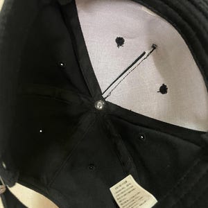 K&ouml;nnte beinhalten: Schwarze Baseballkappe mit wei&szlig;em Innenfutter und einem kleinen Etikett mit japanischem Text. Die Kappe hat einen gebogenen Schirm und einen Knopf oben. Der Innenraum hat drei kleine gestickte Details und eine schwarze Linie.