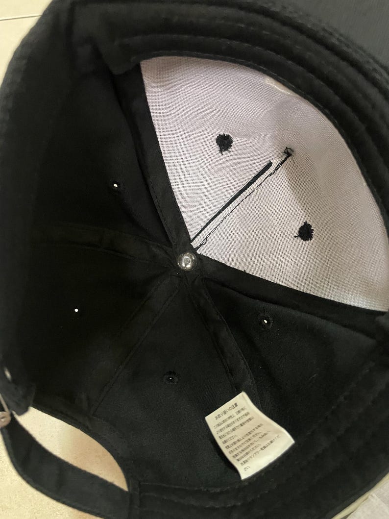 K&ouml;nnte beinhalten: Schwarze Baseballkappe mit wei&szlig;em Innenfutter und einem kleinen Etikett mit japanischem Text. Die Kappe hat einen gebogenen Schirm und einen Knopf oben. Der Innenraum hat drei kleine gestickte Details und eine schwarze Linie.