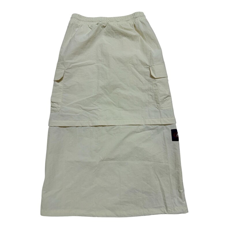 May include: A cream-colored midi skirt featuring an elastic waistband, two side cargo pockets with flaps, and a small rectangular black brand label on the lower right side. The skirt has a layered design with a straight silhouette and smooth fabric.