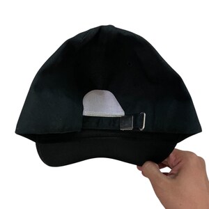 K&ouml;nnte beinhalten: Schwarze Baseballkappe mit gebogenem Schirm und verstellbarem Metallschnallenverschluss. Die R&uuml;ckseite der Kappe hat ein wei&szlig;es Mesh-Panel zur Bel&uuml;ftung. Die Kappe wird von einer Hand gehalten.