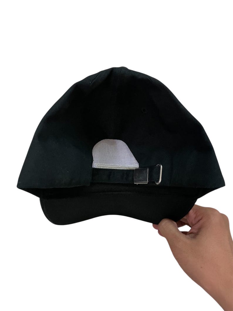 K&ouml;nnte beinhalten: Schwarze Baseballkappe mit gebogenem Schirm und verstellbarem Metallschnallenverschluss. Die R&uuml;ckseite der Kappe hat ein wei&szlig;es Mesh-Panel zur Bel&uuml;ftung. Die Kappe wird von einer Hand gehalten.