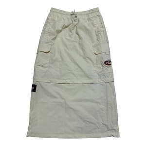 May include: A cream-colored midi skirt featuring an elastic waistband with a drawstring, two side cargo pockets, and a Fila logo patch on the left pocket. The skirt has a horizontal seam across the middle and a small Fila brand tag on the lower side.