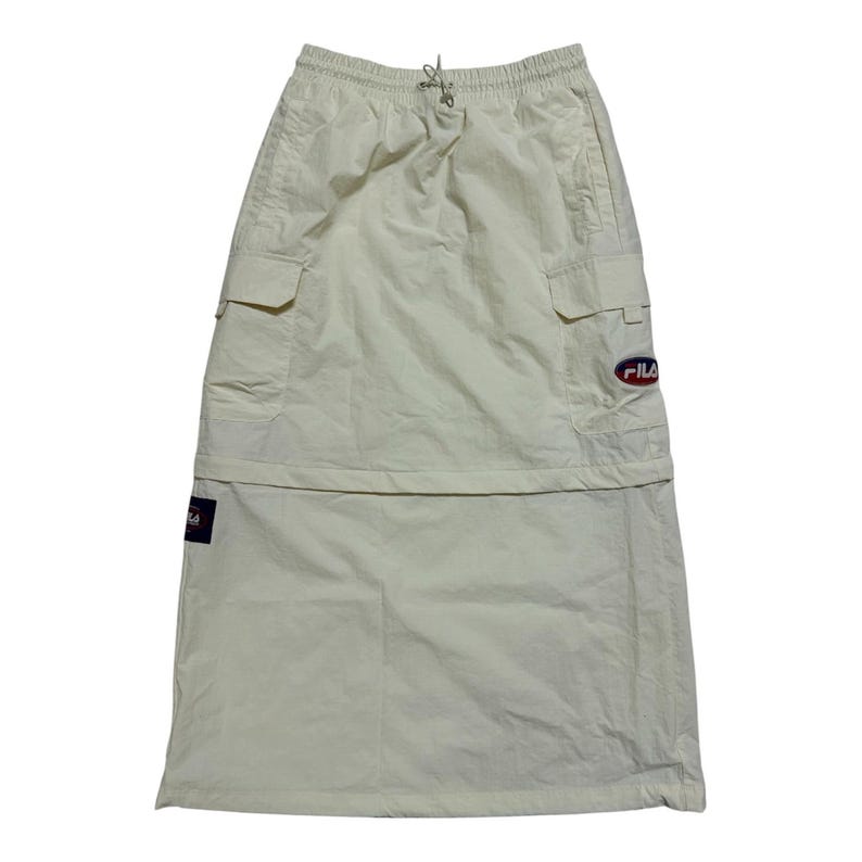 May include: A cream-colored midi skirt featuring an elastic waistband with a drawstring, two side cargo pockets, and a Fila logo patch on the left pocket. The skirt has a horizontal seam across the middle and a small Fila brand tag on the lower side.