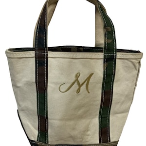 May include: Cream-colored canvas tote bag with camouflage accents on the handles and trim. The bag features an embroidered gold letter "M" on the front. The handles are sturdy, and the bag appears to be well-constructed for carrying items.