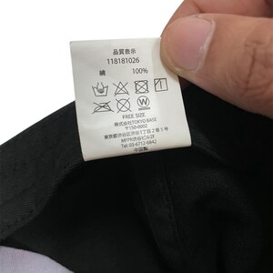 K&ouml;nnte beinhalten: Nahaufnahme eines Etiketts mit japanischem Text und Pflegesymbolen. Auf dem Etikett steht "100%" und "FREE SIZE". Das Etikett wird &uuml;ber einem schwarzen Stoff gehalten.