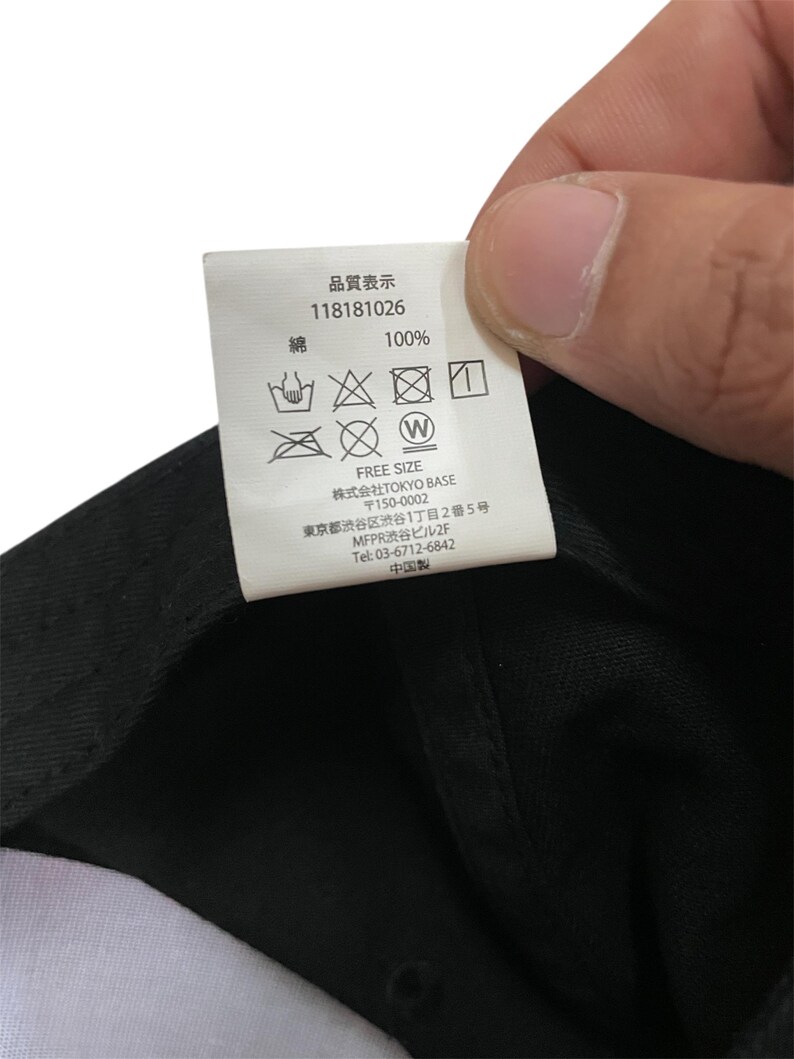 K&ouml;nnte beinhalten: Nahaufnahme eines Etiketts mit japanischem Text und Pflegesymbolen. Auf dem Etikett steht "100%" und "FREE SIZE". Das Etikett wird &uuml;ber einem schwarzen Stoff gehalten.