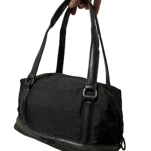May include: Black handbag with leather handles and trim. The bag has a rectangular shape with a rounded top and a fabric body. The handles are attached to the bag with silver-colored metal rings.