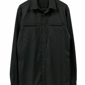 Pode incluir: Uma camisa preta de manga comprida com dois bolsos no peito com zíper e uma frente abotoada. A camisa está pendurada em um cabide de madeira. O logotipo da Prada é visível.